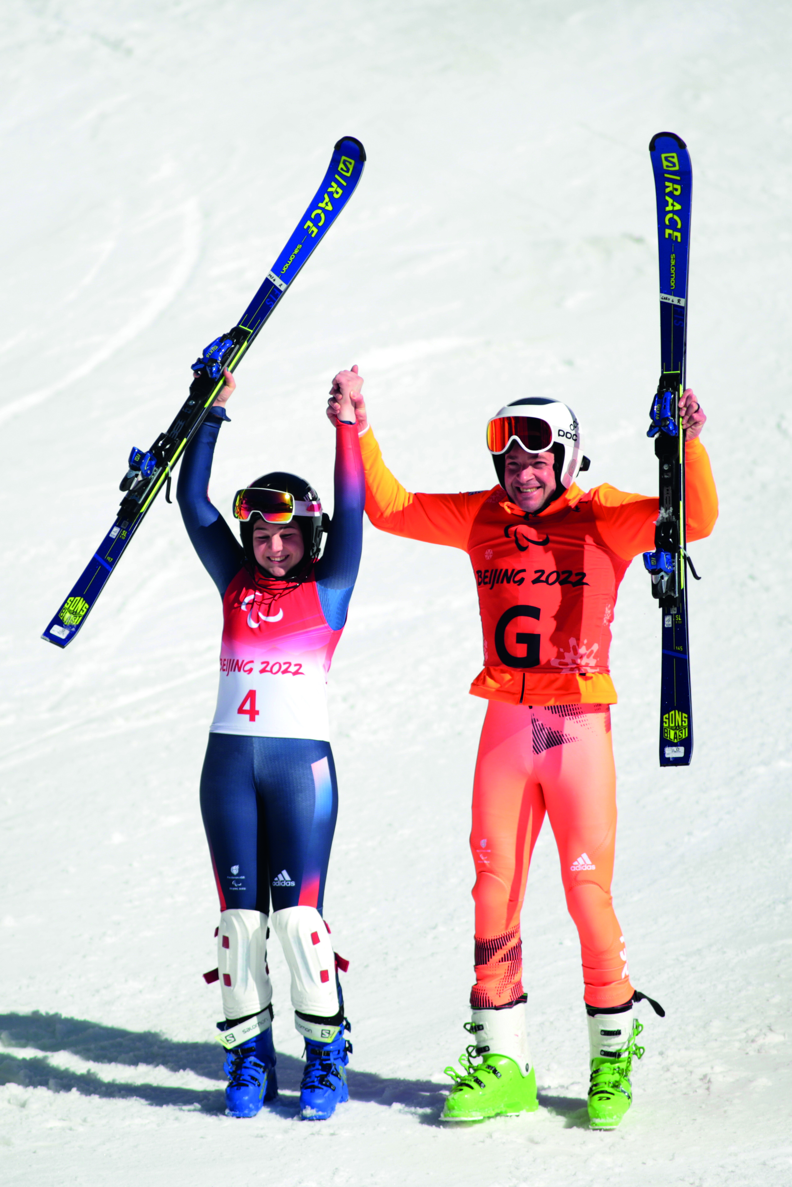 Gary and Menna hold their skis in the air, on the slopes.
